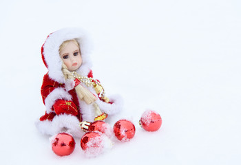 Doll in a red coat sitting in the snow around the red balls.
