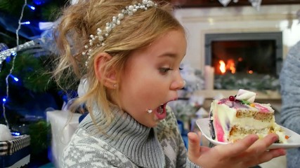 girl with pleasure eating a large piece of cake at The Christmas tree, Family by the fireplace eating a dessert with tea, mom and daughter enjoying spending time together, in winter - Powered by Adobe