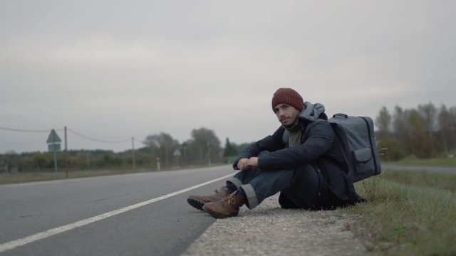 Casualy Dressed Traveler Sitting by the Highway, Weather is Cloudy. Shot on RED Cinema Camera in 4K (UHD). 