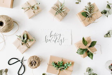 calligraphy words happy holidays and beauty arrangement frame of craft boxes and green branches on white background. flat lay, top view