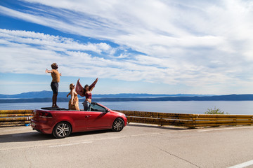 Group of happy young people standing in the red convertible car and waving. - obrazy, fototapety, plakaty