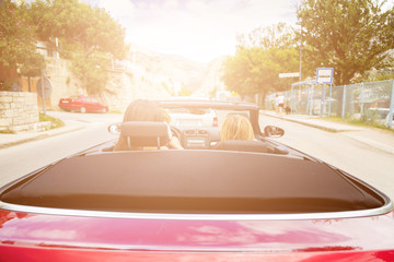 Back view of two girls driving red convertible car. Sun effect applied.