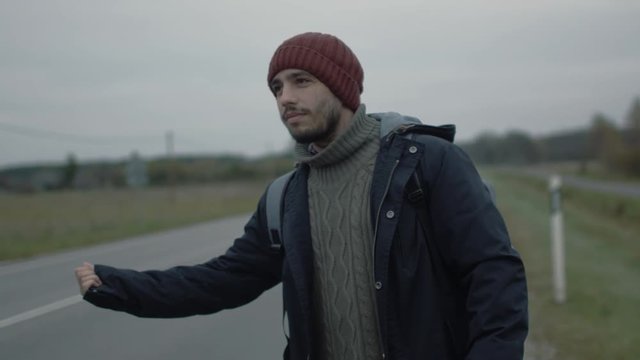 Casually Dressed Traveler standing by the Highway and Thumbing at a Cloudy Day