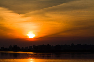 Sunrise over the surface water of lake with floating birds and curtain clouds in the red sky.