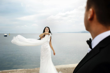 Look over groom's shoulder at bride holding her veil against the