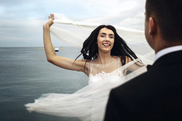 Happy brunette bride whirls her veil standing before groom on th