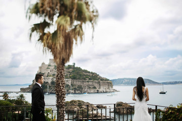 Groom walks to long-haired bride watching the yachts from balcon
