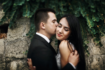 Fototapeta premium Bride looks down enjoying groom's kiss before the wall with gree