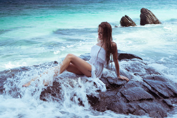 Sensual brunette woman in white dress with wet hair sitting on the rock over beautiful sea background