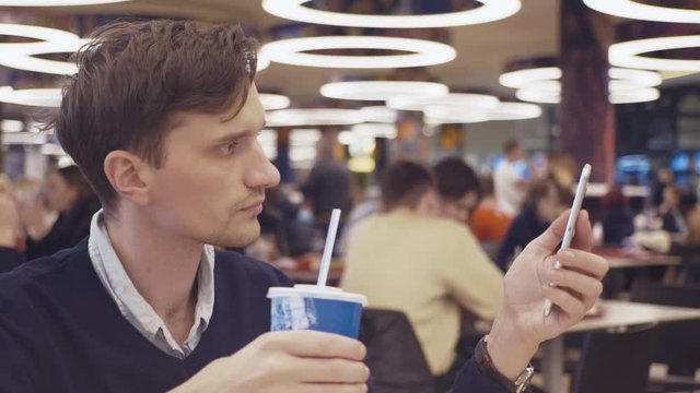 Young men using smartphone and drinking coke at food court in shopping mall. Portrait