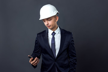 Handsome young engineer in a helmet  looking in phone  standing on gray background