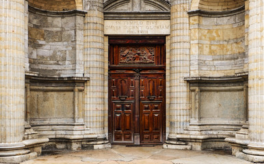 Fototapeta na wymiar Heavy wooden double door outside of an old church. Circle wall with square stones and pillars. Latin inscriptions on the sides and the arch. Religious symbolic relief on top. Rectangle door frame.