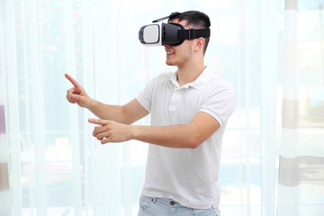 Young man wearing virtual reality glasses and standing near window in a room