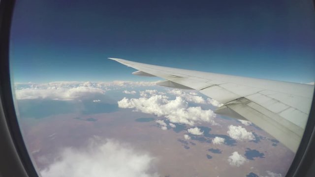 In-flight Airliner Wings Over Moving Clouds Seen from passenger Window with Arid Desert Land Underneath on a Sunny Day with a Blue Sky