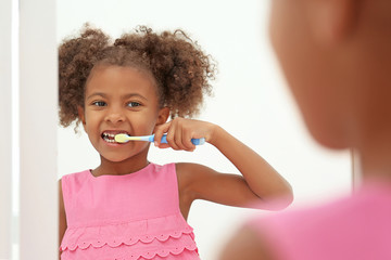 Cute African American girl brushing teeth and looking in mirror in bathroom
