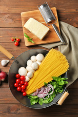 Pan with ingredients for tasty pasta, grater, napkin and kitchen board on wooden table, top view