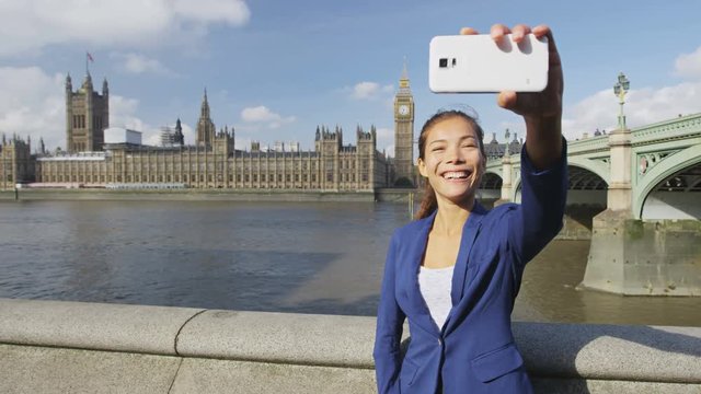 Businesswoman taking selfie using phone in London on business travel. Business woman smiling at camera with Thames river, Big Ben and Westminster bridge background. Asian woman using smartphone.