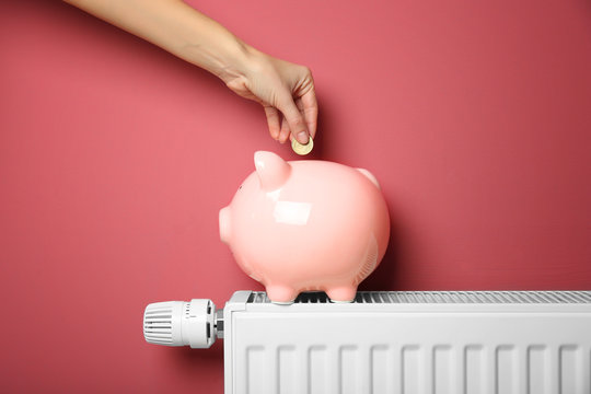 Savings concept. Female hand putting coin into piggy bank which standing on heating radiator with temperature regulator on pink background