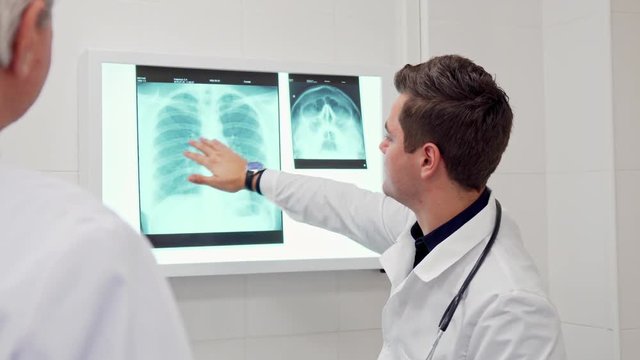 Young male doctor showing something on x-ray to his elder colleague. Senior gray man in white coat standing backwards the camera. Brunette caucasian man moving his hand on the x-ray image