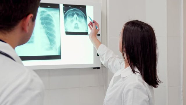 Mature female doctor pointing pen on the x-ray image of human head. Young brunette man in white coat standing backwards near his female coworker. Attractive caucasian female medic consulting with her