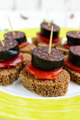 Sandwiches with black rye bread in the shape of a heart, blood sausage (Morcillo) and pieces of sweet pepper on skewers and tomato sauce, on a white wooden background. Close up