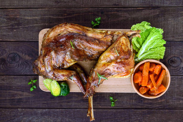 Roast whole rabbit on a wooden board with baked carrots and Brussels sprouts on a dark background. A festive meal. The top view.