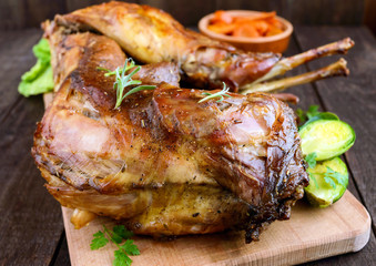 Roast whole rabbit on a wooden board with baked carrots and Brussels sprouts on a dark background. A festive meal.