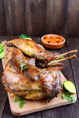 Roast whole rabbit on a wooden board with baked carrots and Brussels sprouts on a dark background. A festive meal.