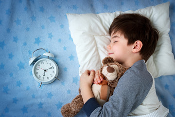 six years old child sleeping in bed on pillow with alarm clock and a teddy bear