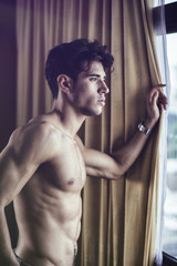 Sexy handsome young man standing shirtless in his bedroom next to window curtains