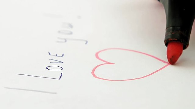 Female hand draws red heart valentine under I love you! phrase on the sheet of white paper. Macro close-up shot.