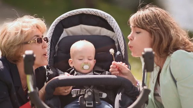 Baby in a stroller outdoors. Next, mother and grandmother blow on him dandelions.