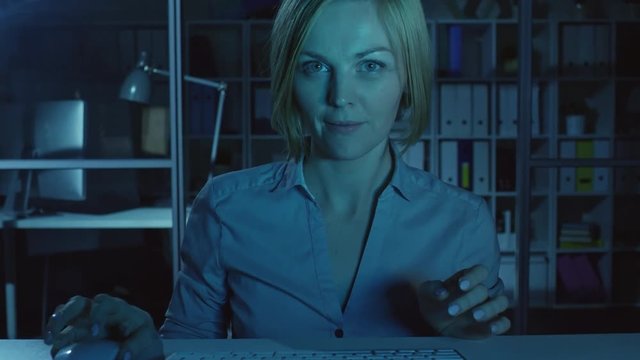 Joyous woman sitting at desk in the dark office, typing on keyboard, reading something on computer screen and smiling