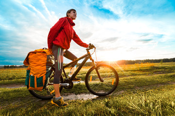 Young lady hiker with loaded bicycle standing on a wet rural road in the meadow