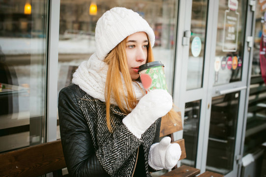 woman in white scarf and hat drinks coffee. cute young blonde girl sitting on a bench near the coffee shop, drinking takeaway coffee, holding Christmas paper cup in his hands. cup to his mouth