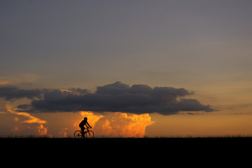 Fototapeta premium Silhouette image of a cyclist (young man or woman) on the road with sunset and clouds in the background
