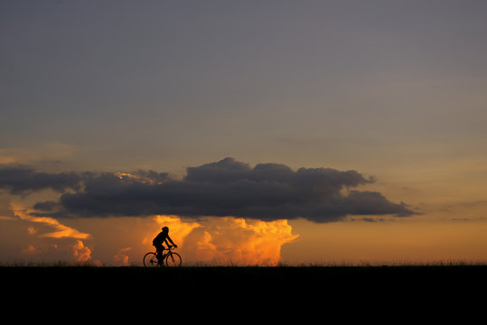 Silhouette image of a cyclist (young man or woman) on the road with sunset and clouds in the background