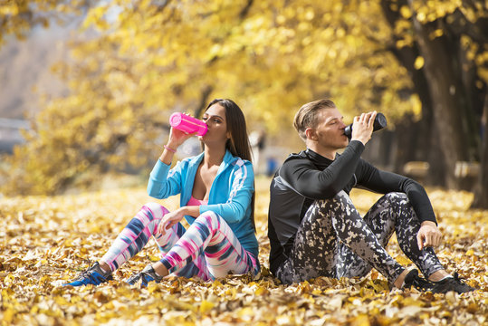 Beautiful young couple resting and rehydration after successful training in the park. Autumn environment.