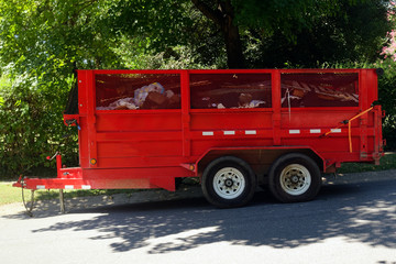 Red utility trailer parked in shade filled with trash and debris. Horizontal.