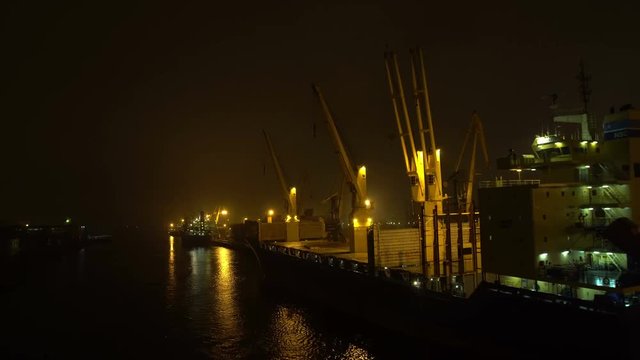 Ship leaving harbor sailing along the jetty pier with many crane spreaders and containers at the night
