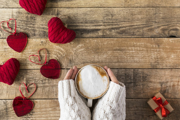 cup of hot cappiccino on wooden table with decorative glass and knitted hearts around