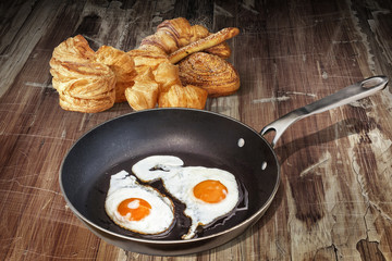Fried Eggs In Old Teflon Frying Pan With Bunch Of Croissant Puff Pastry Sprinkled With Sesame Seeds Set On Old Cracked Flaky Wooden Table