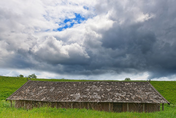 Fototapeta na wymiar Vintage squat wooden shed on field under sky with stormy clouds.