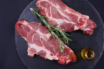 Raw pork neck meat on black background, rosemary, olive oil. Cooking background, healthy eating concept.