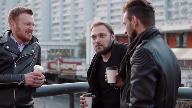 Three handsome young men with beards talk, smile and have coffee on the go near a bridge railing in the city. Slow mo