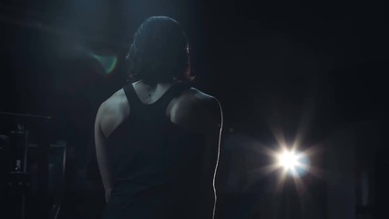 Strong toned young woman in a darkened gym standing with her back to the camera and muscles defined by a small light in the distance
