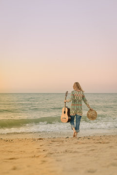 Elegant woman walking on the beach with guitar back view, outdoor background