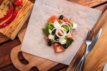 Concept: restaurant menus, healthy eating, homemade, gourmands, gluttony. Greek salad served on paper with ingredients on weathered wooden background. Top-down view.