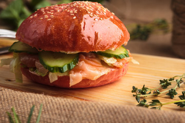 Concept: restaurant menus, healthy eating, homemade, gourmands, gluttony. Trendy glossy burger with salmon in pink bun with ingredients on messy vintage wooden background.