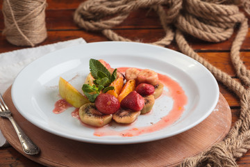 Concept: restaurant menus, healthy eating, homemade, gourmands, gluttony. White plate of fruit flambe. Apple, orange, strawberry, kiwi flambeed in cognac. On weathered wooden table.